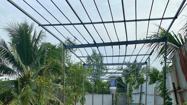 A covered pathway with tiled flooring, surrounded by lush greenery and a metal fence.