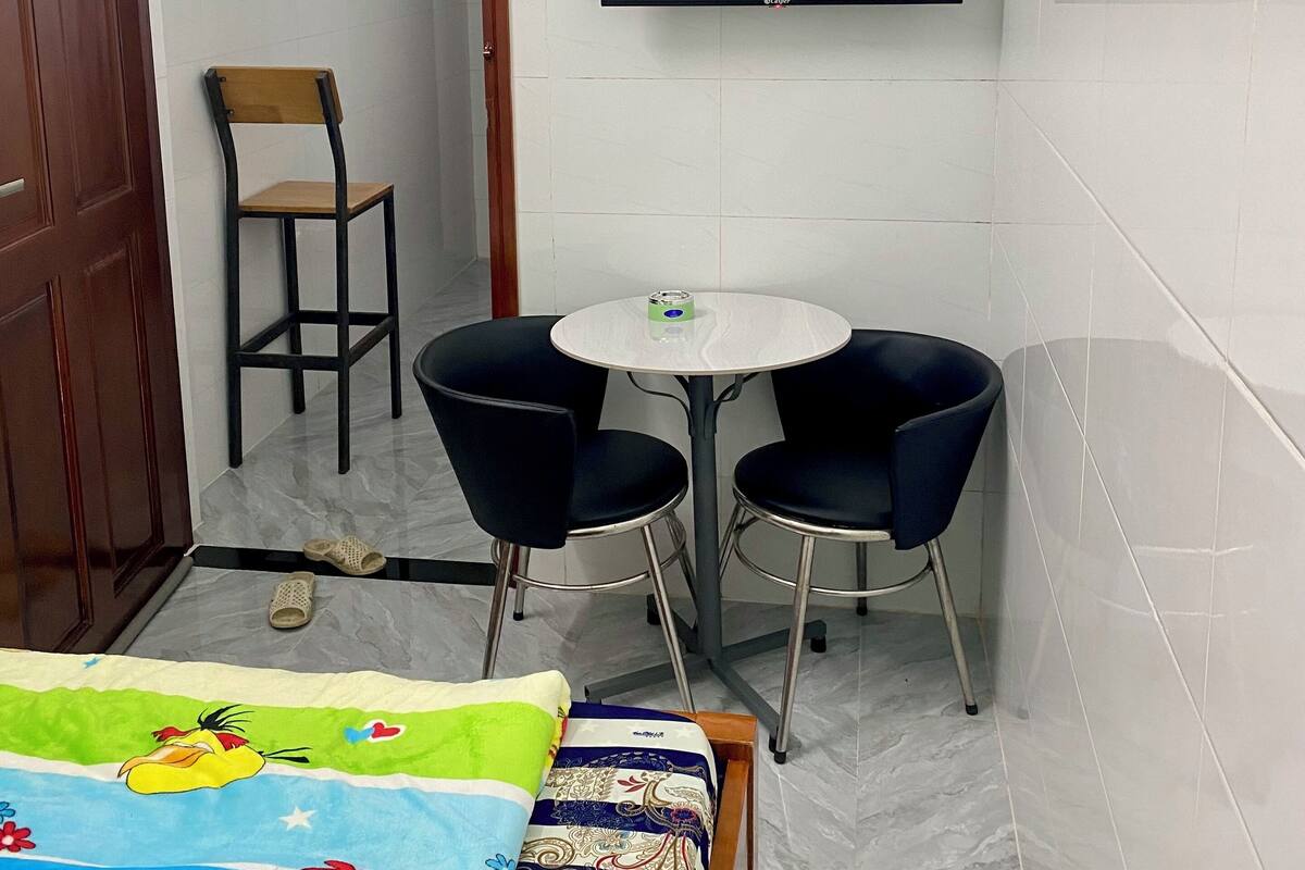 Una habitación pequeña con una cama, televisión instalada en la pared, dos sillas, una mesa y una puerta.