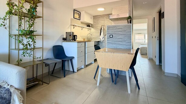 A modern kitchen with a coffee machine, a dining table with chairs, a blue chair, and a plant on a metal shelf. - T2 Apartment with Terrace in Port-Vendres Center (Port-Vendres)