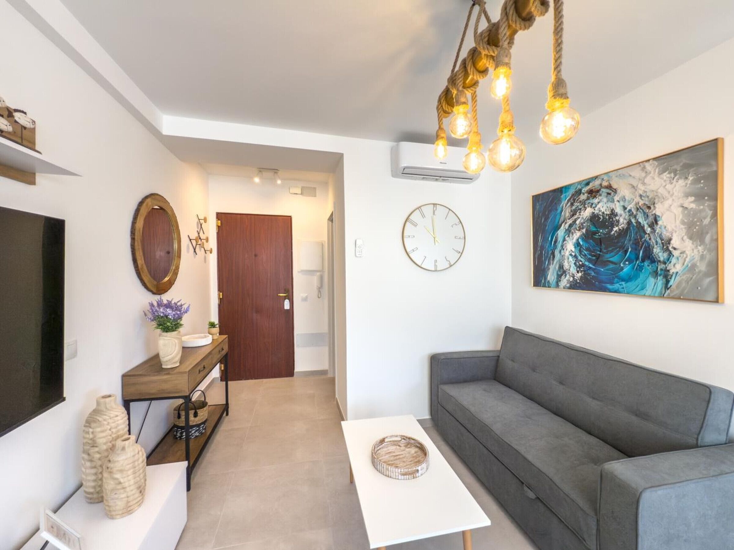 Un saló modern amb un sofà gris, una televisió de pantalla plana, una taula auxiliar de fusta amb objectes decoratius, una taula de cafè blanca i un gran quadre abstracte a la paret.