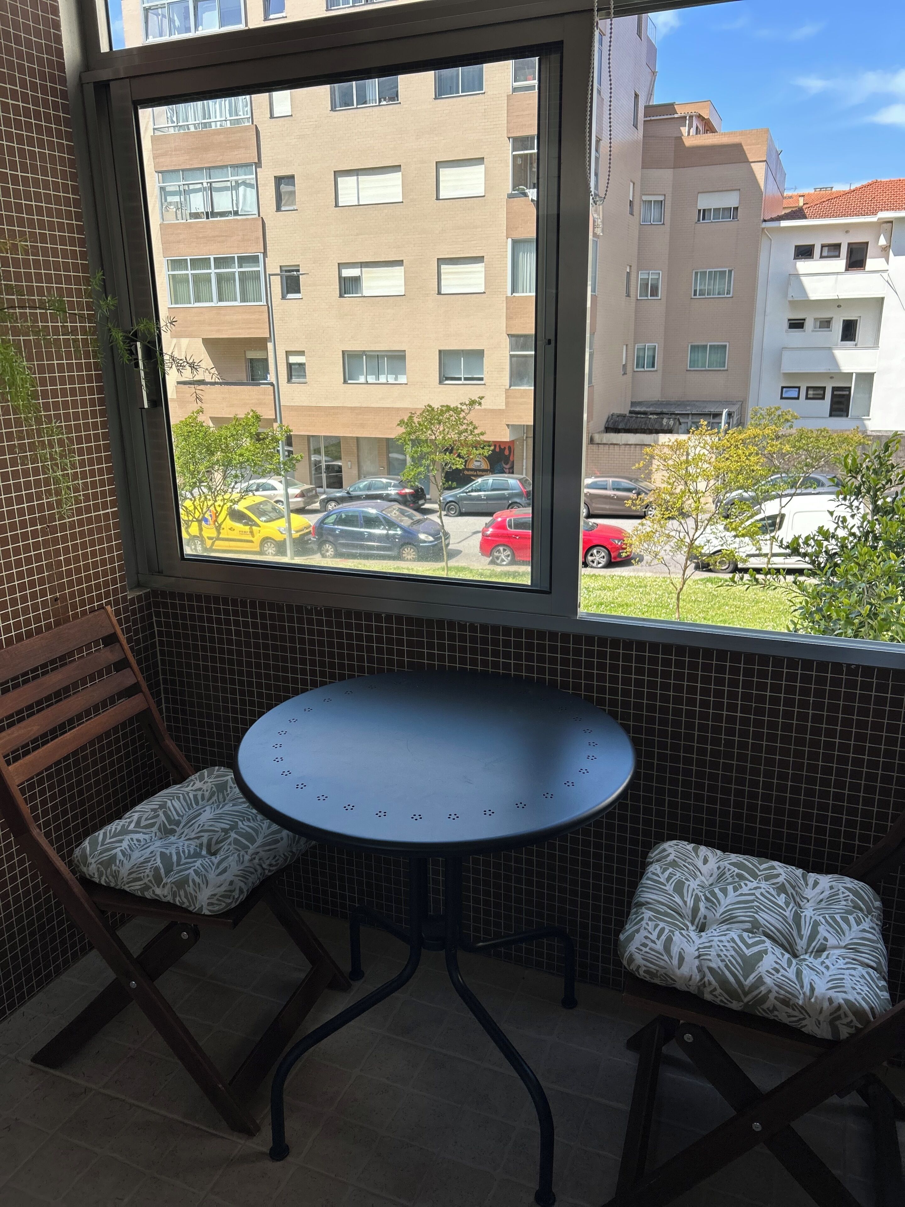 Un balcon avec une table ronde et deux chaises, un mur recouvert de carreaux et une vue sur un immeuble et des voitures garées.