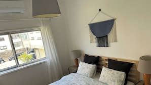A bedroom with a bed, bedside tables, a hanging macrame wall hanging, a window with curtains, and an air conditioning unit. - Casa da Praceta (Porto)