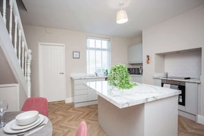 A modern kitchen with a marble island, a dining area with a pink chair, and a staircase with white railings. - Lydia Terrace | Parking | Wi-Fi (County Durham)