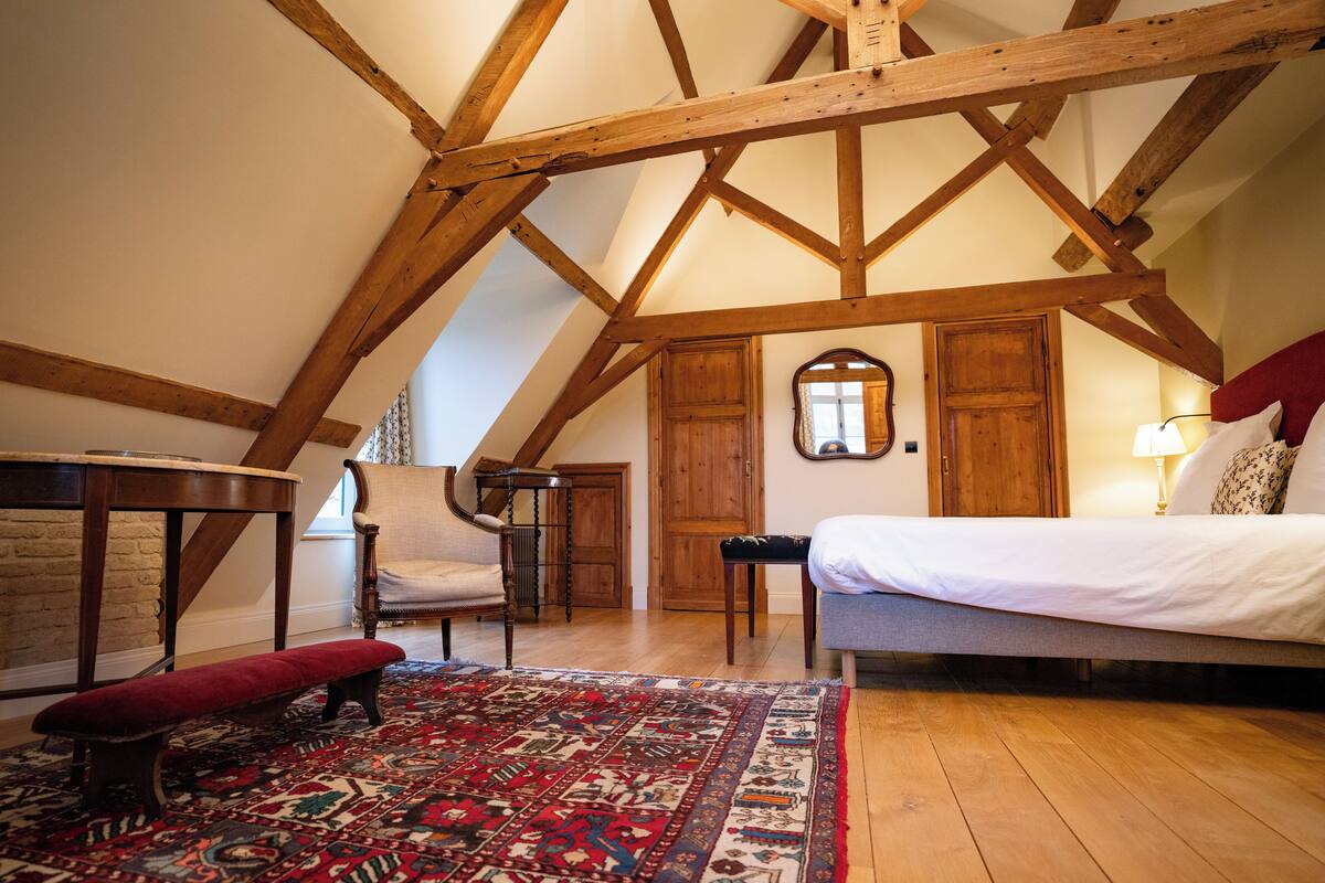 A bedroom with a sloped ceiling, wooden beams, a bed, a chair, a bench, and a patterned rug.