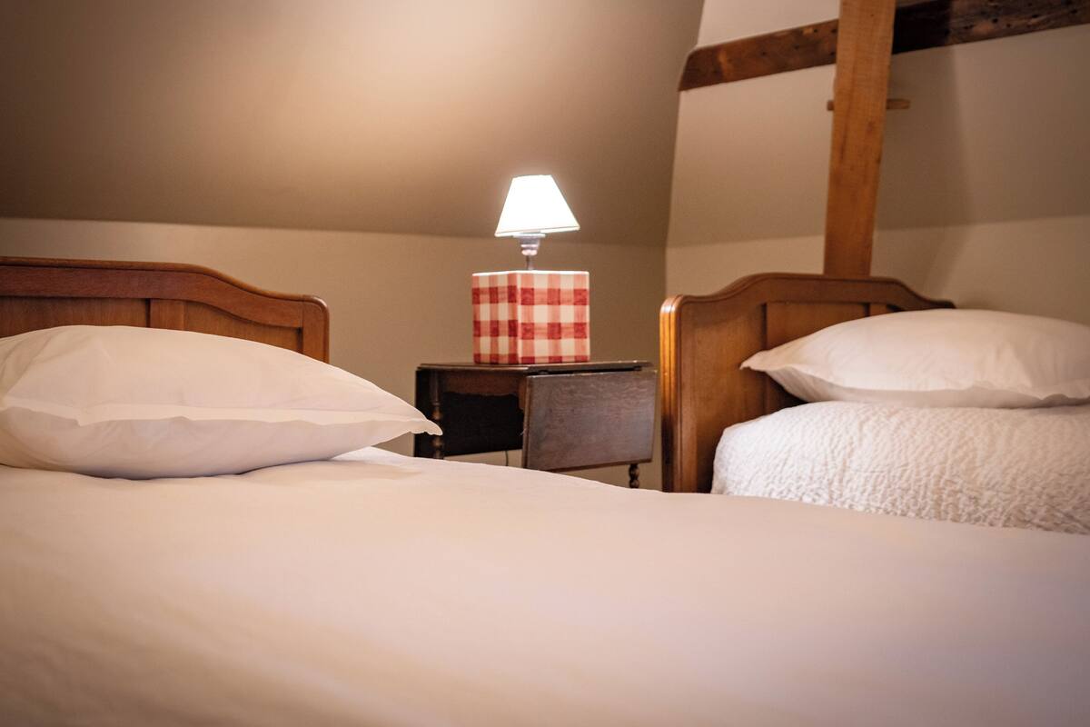 A hotel room with two beds, each with white bedding and pillows. There is a small table with a red and white checkered tablecloth and a lamp on it. The room has wooden beams on the ceiling and a simple, clean design.