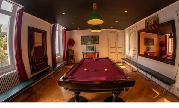 Une table de billard avec des billes, une queue de billard et un luminaire vert suspendu dans une pièce au plancher de bois et ornée d’un grand miroir au mur.