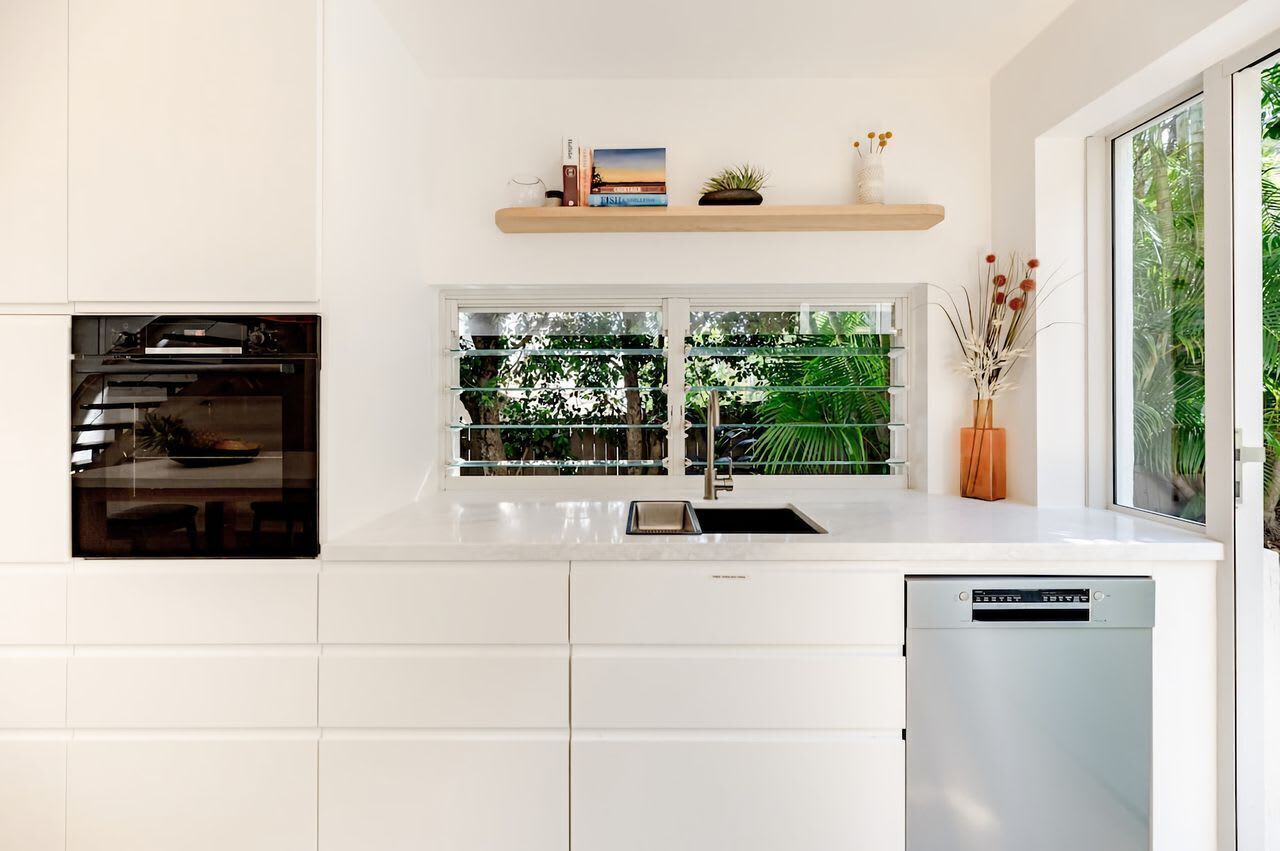 A modern kitchen with a built-in oven, a window with a view of greenery, and a dishwasher.