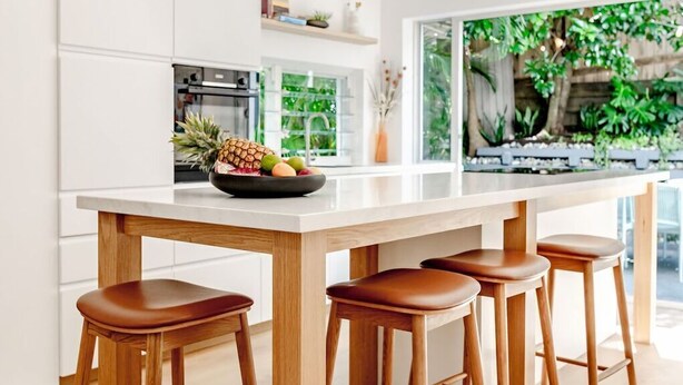 A modern kitchen with a central island, wooden stools, and a hanging light fixture.