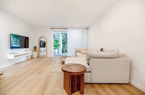 A modern living room with a large flat-screen TV, a beige sofa, a wooden coffee table, and a view of greenery through a glass door.