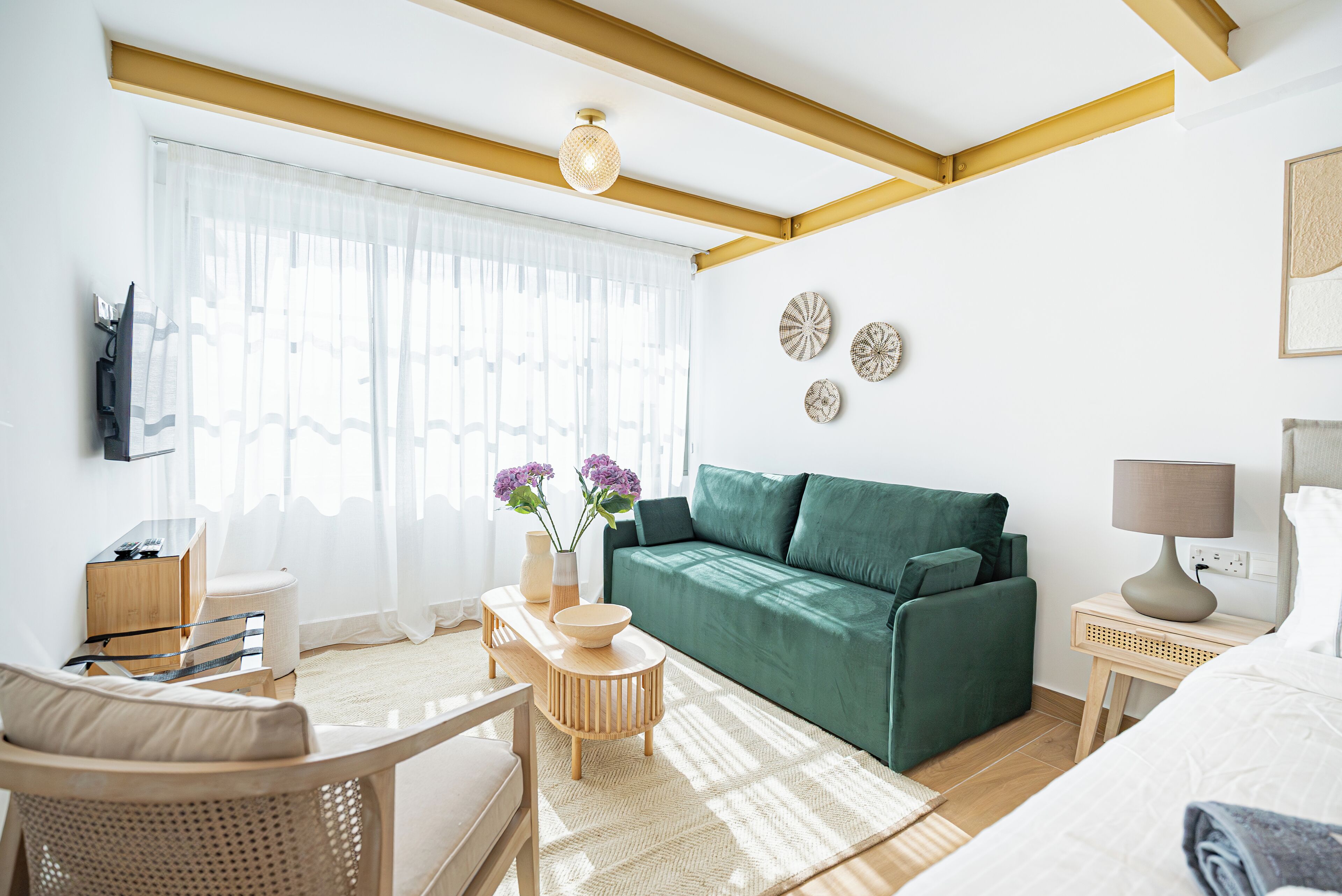 A modern hotel room with a green sofa, a wooden chair, a small table with a bowl, and a lamp on a bedside table.