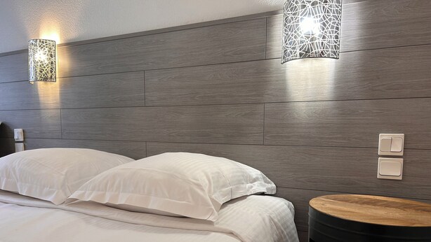 A modern hotel room with a wooden headboard, two bedside lamps, a round wooden side table, and a neatly made bed with white linens. - Hotel Le Lacuzon (Moirans-en-Montagne)