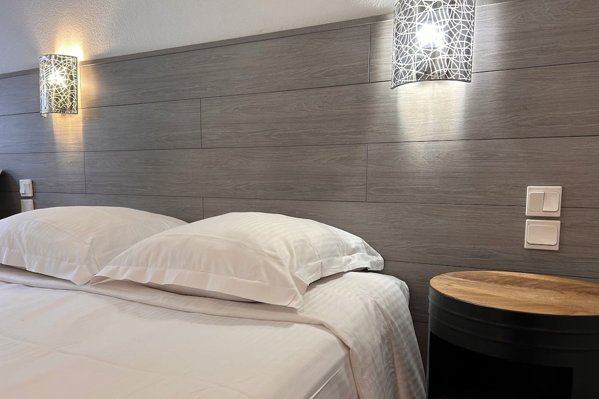 A modern hotel room with a wooden headboard, two bedside lamps, a round wooden side table, and a neatly made bed with white linens.