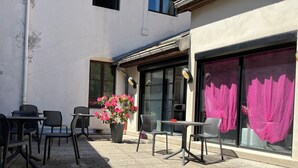 An outdoor seating area with tables and chairs, a potted plant with flowers, and a building with large windows and a door. - Hotel Le Lacuzon (Moirans-en-Montagne)