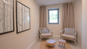 Ein Zimmer mit zwei Stühlen, einem kleinen runden Tisch, einem Fenster mit Vorhängen und gerahmten Bildern an der Wand.