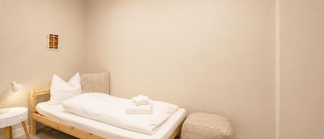 A single bed with a wooden frame, white bedding, a bedside table with a lamp, a small stool, and a wall-mounted shelf.