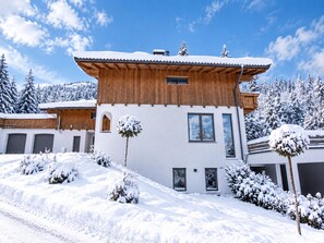 Un bâtiment moderne à deux étages, avec un étage supérieur en bois et un étage inférieur blanc, entouré d’arbres couverts de neige et sous un ciel dégagé.