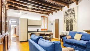 A modern living room with a blue sofa set, a kitchen in the background, and a large abstract painting on the wall. - ***Newly Restored*** Spanish Steps Cosy apartment (Roma)