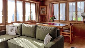 A cozy cabin interior with a sofa, wooden dining table, and large windows with curtains. - Stunning  villa for 3 people with TV, patio, pets allowed and panoramic view (Habkern)