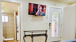 A bedroom with a bed, a TV mounted on the wall, a bench, and a door with a window. - The Studio on Polk - Cozy Downtown Studio! (Brownsville)