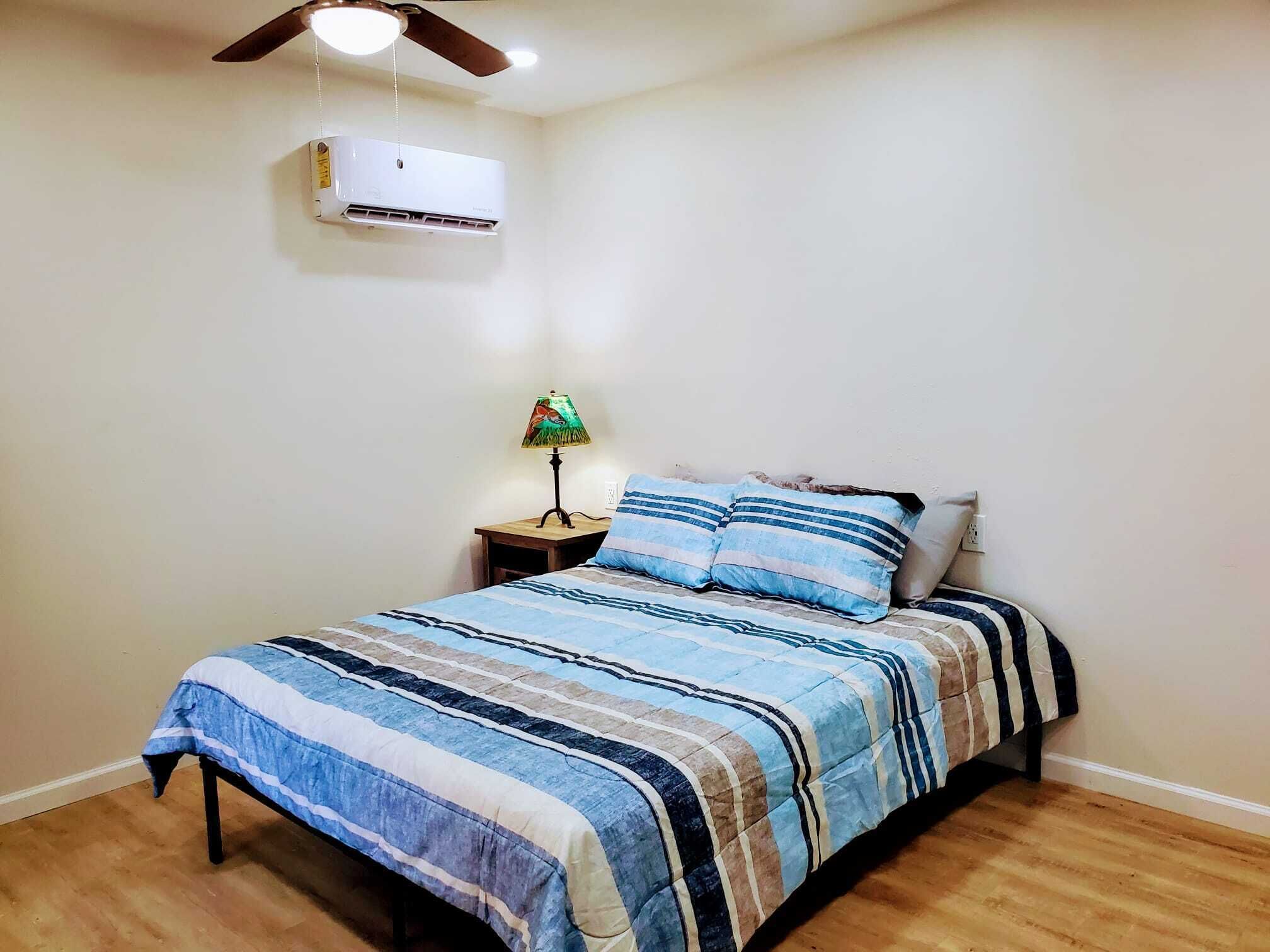 Un dormitorio con cama, mesita de noche, lámpara, aire acondicionado y ventilador de techo.