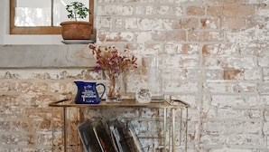 A rustic brick wall, a window with a potted plant, a glass vase with dried flowers, a blue bag, and a metal sculpture on a metal table. - King St Cottage: Central and Stylish (Hobart City)