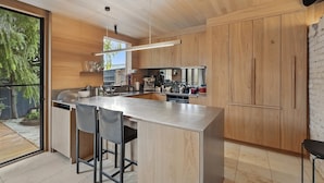 A modern kitchen with wooden cabinets, a large island with bar stools, and a view of the outdoors. - King St Cottage: Central and Stylish (Hobart City)