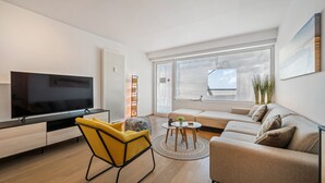 A modern living room with a flat-screen TV, a sofa, a yellow armchair, and a small round table with a plant.