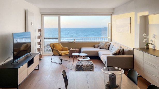 A modern living room with a large TV, a sofa, a dining table with chairs, and a view of the sea through large windows.
