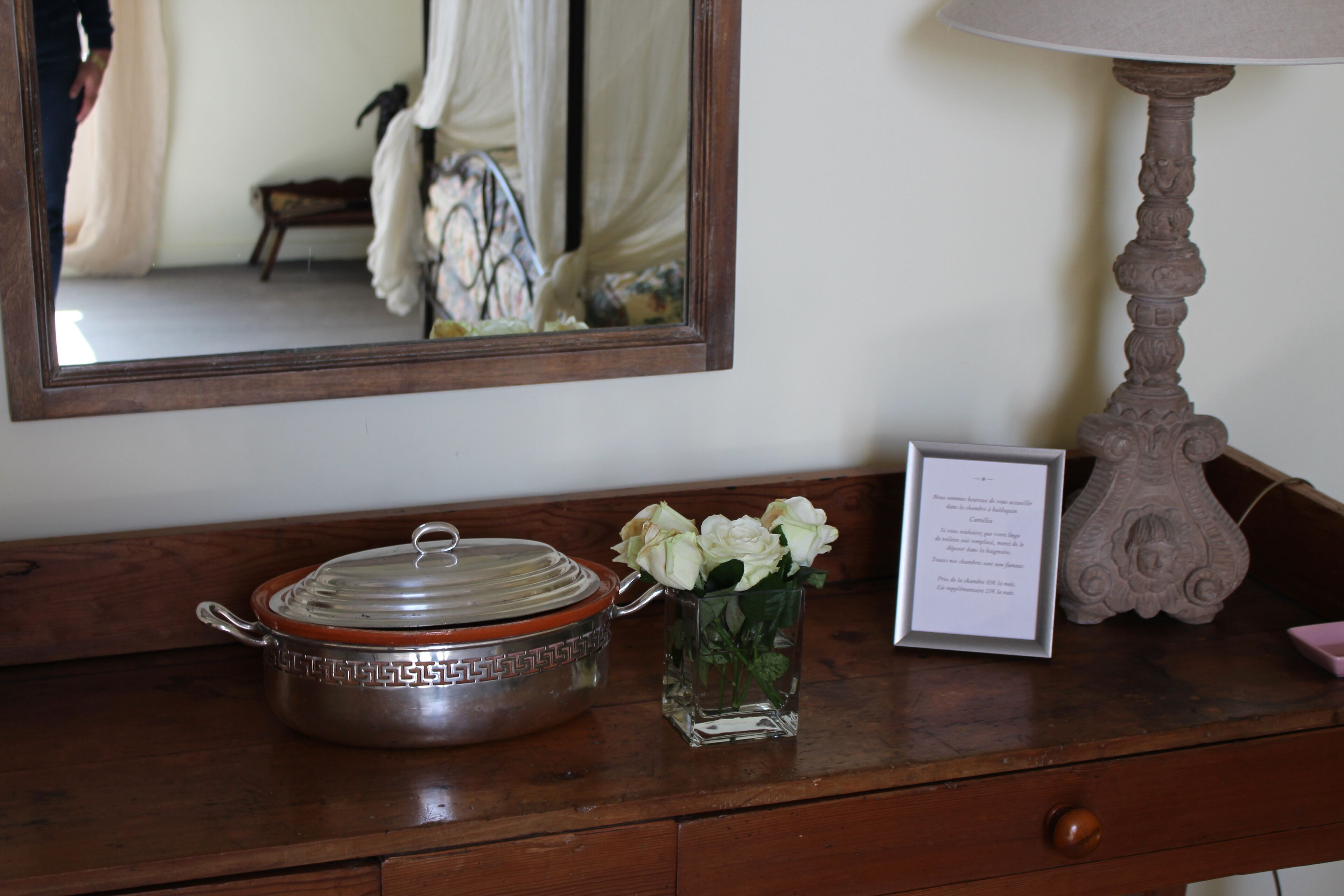 A wooden dresser with a metal pot, a vase with white flowers, a framed note, and a lamp.