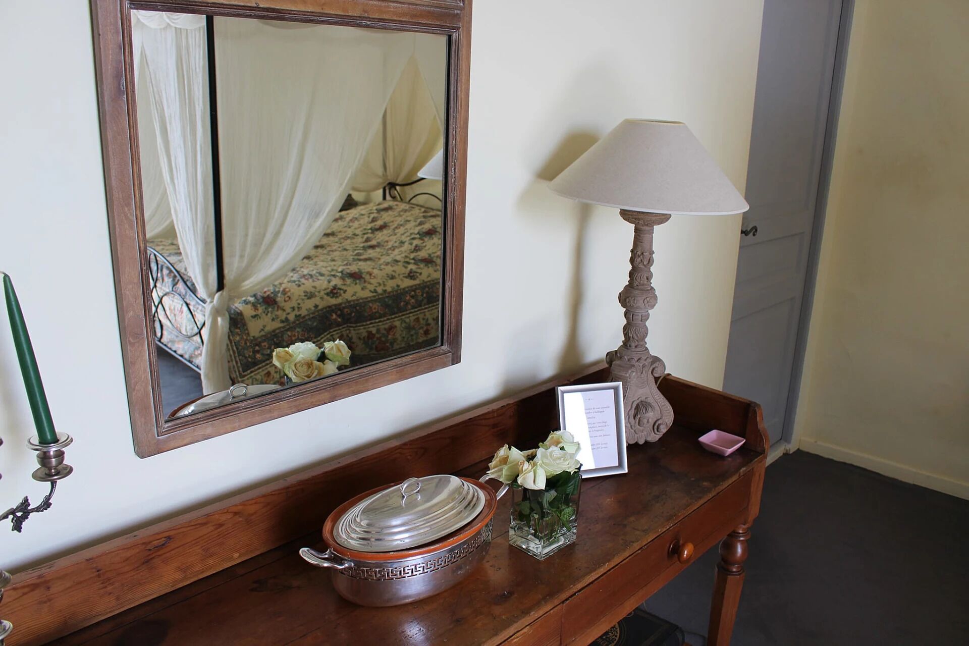 A wooden console table with a mirror, a lamp, a vase with flowers, and a framed menu.