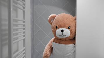 Un baño con ducha de azulejos blancos, un toallero blanco y un gran oso de peluche.