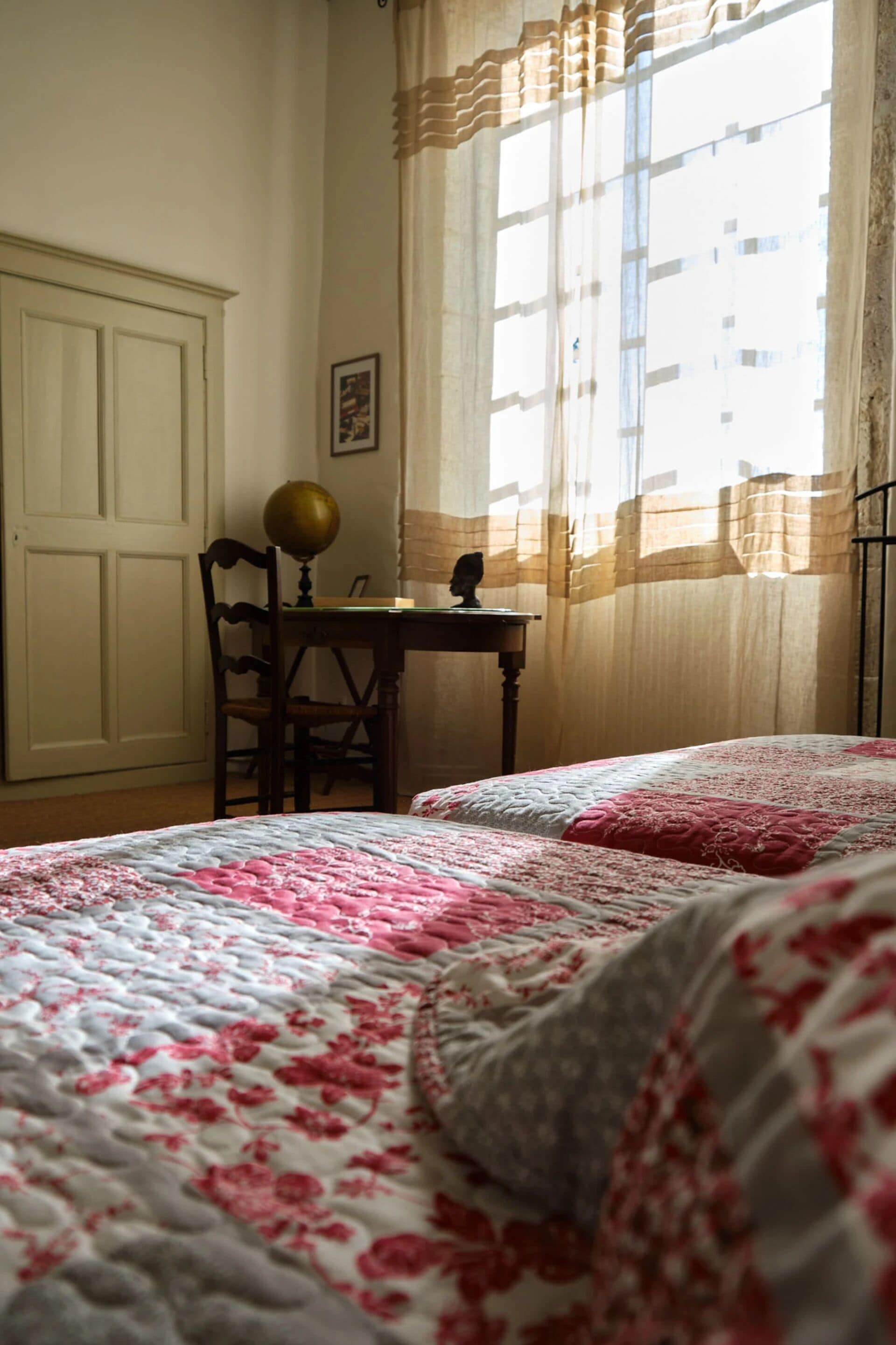A bedroom with two beds, a desk with a globe, a chair, and a window with curtains.