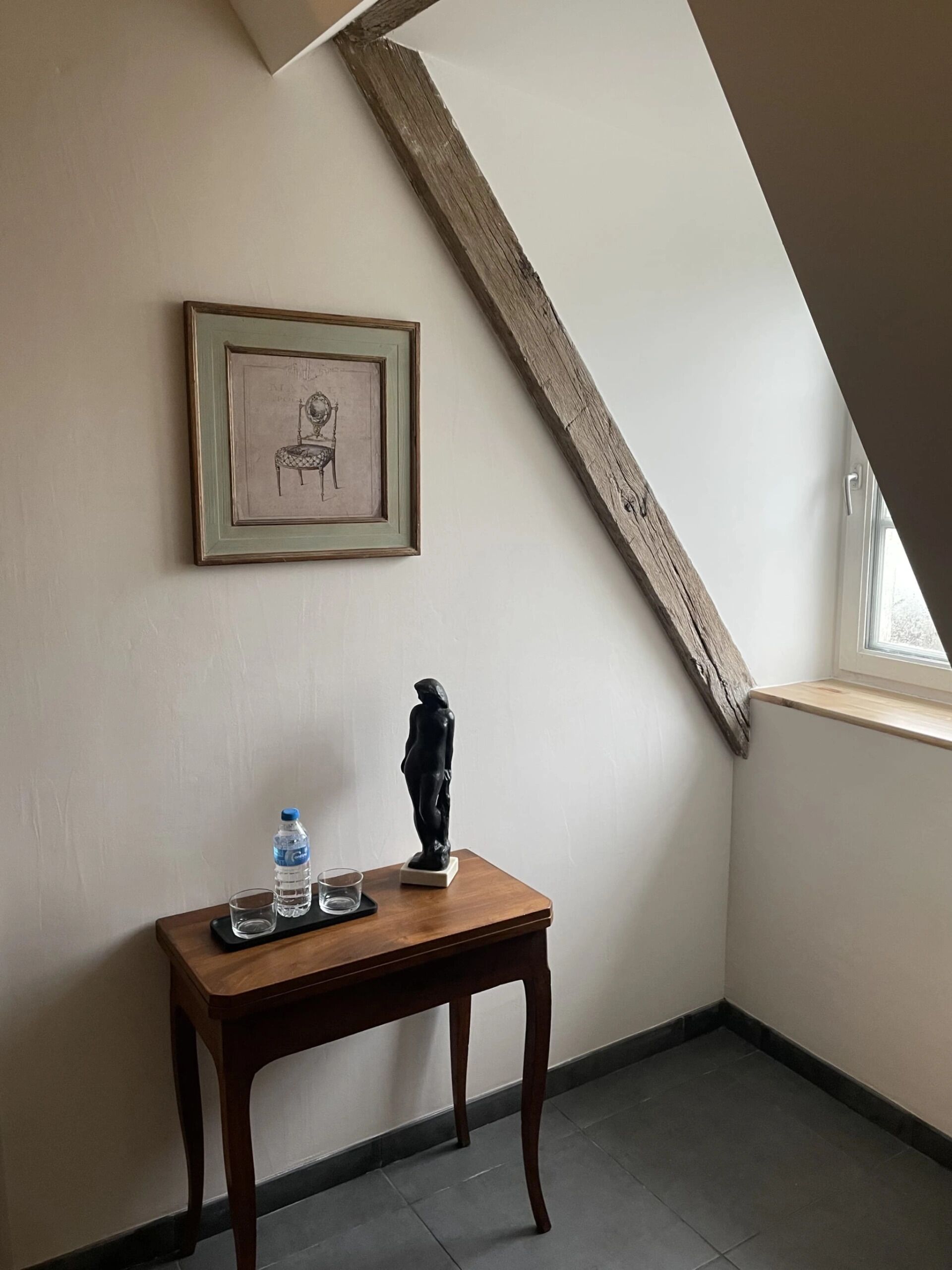 Une petite pièce avec un plafond en pente, un tableau encadré au mur, une statue posée sur une table en bois et une bouteille d’eau.