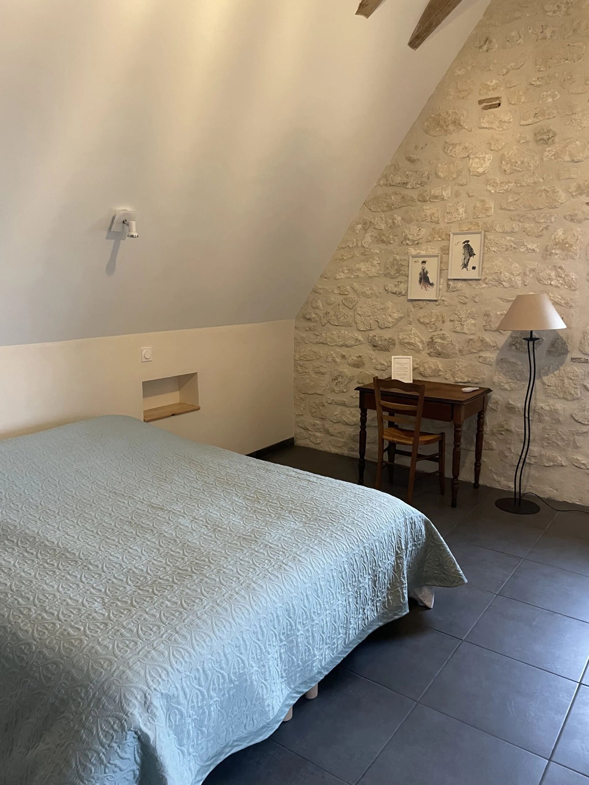Een slaapkamer met een bed, een bureau, een stoel, een lamp en een stenen muur.