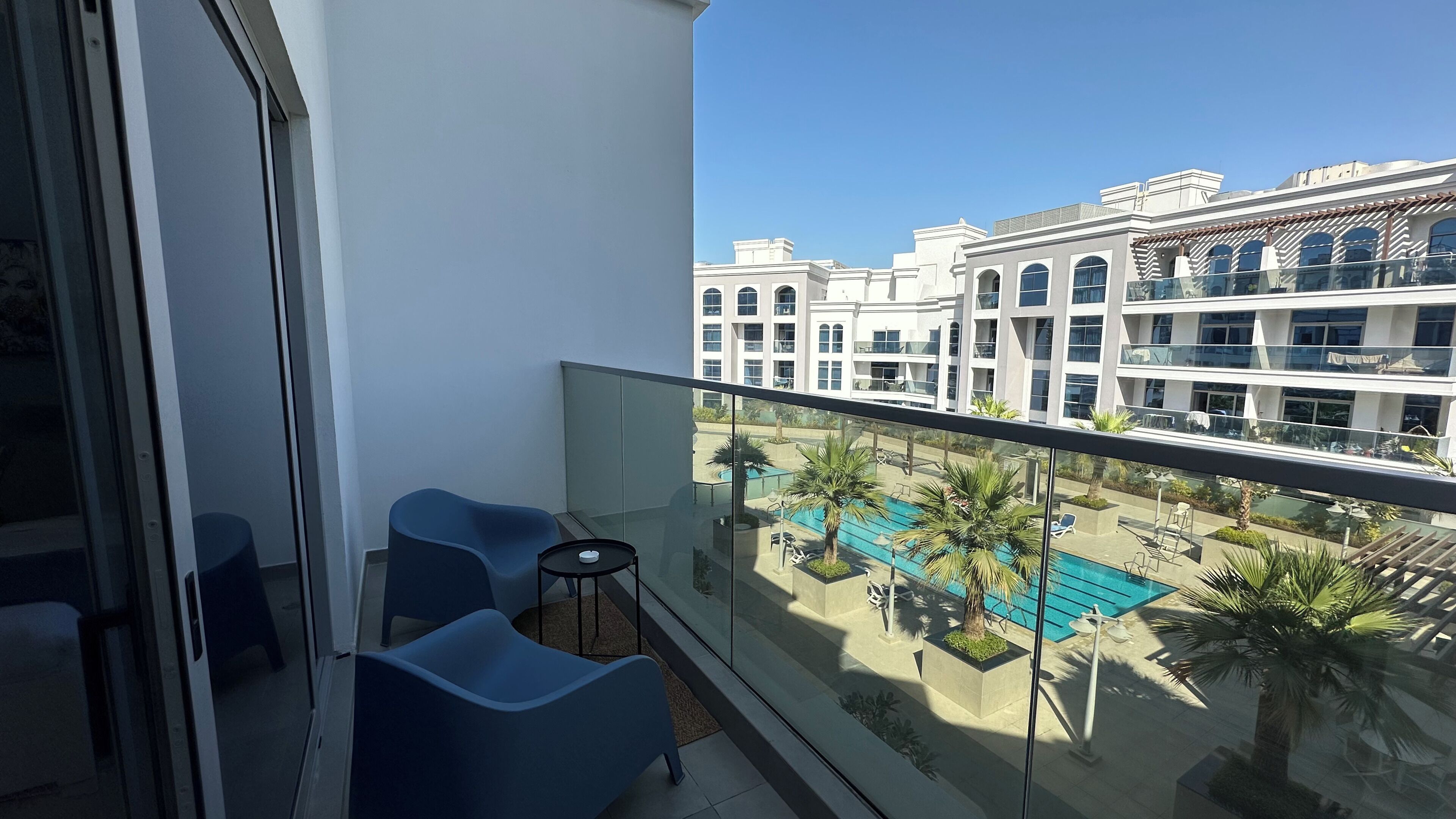 A balcony with two blue chairs and a small table, overlooking a swimming pool and a building with multiple balconies.