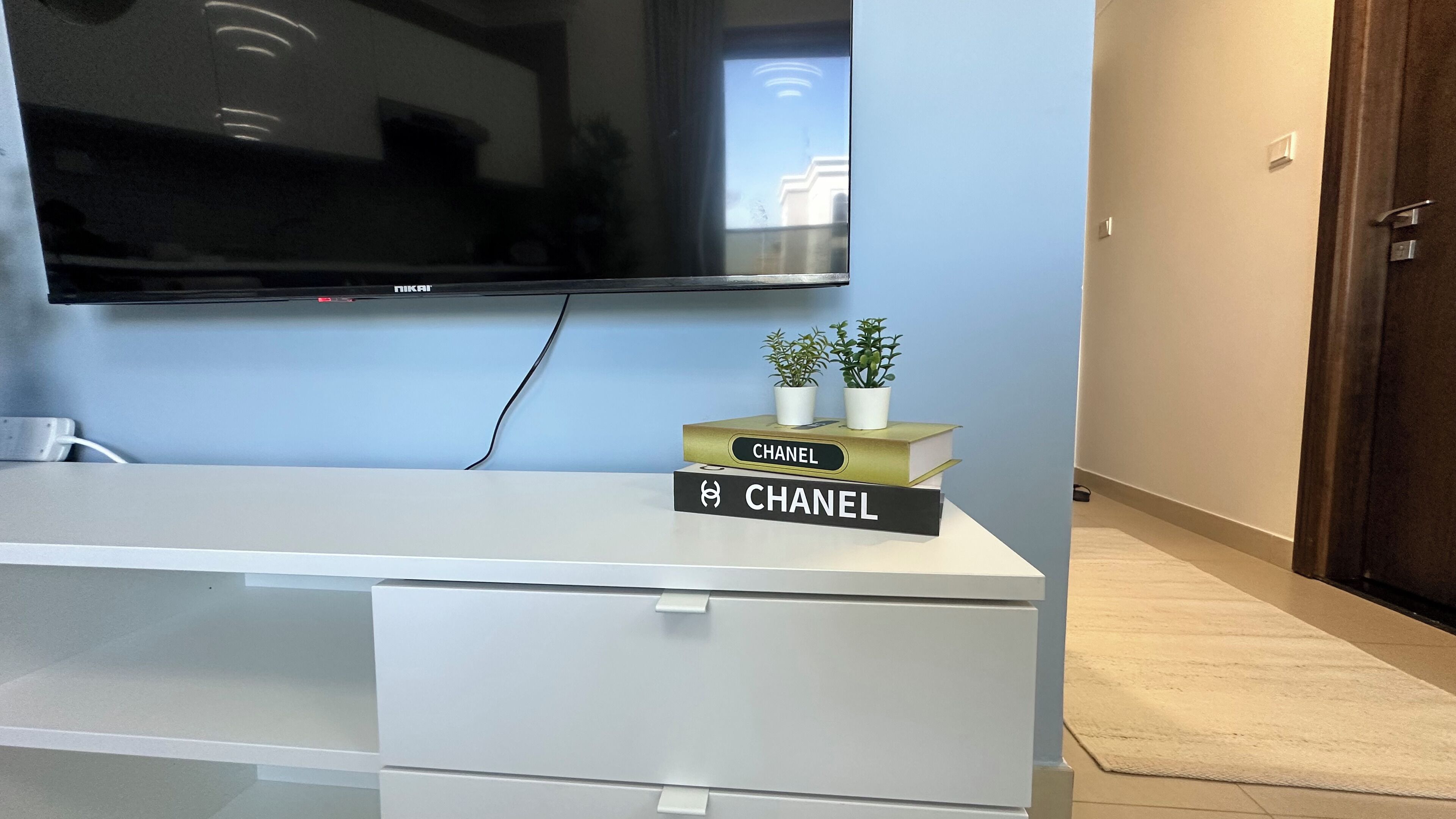 A modern living room with a flat-screen TV mounted on the wall, a white entertainment unit with two books titled 'CHANEL', and a small potted plant on top.