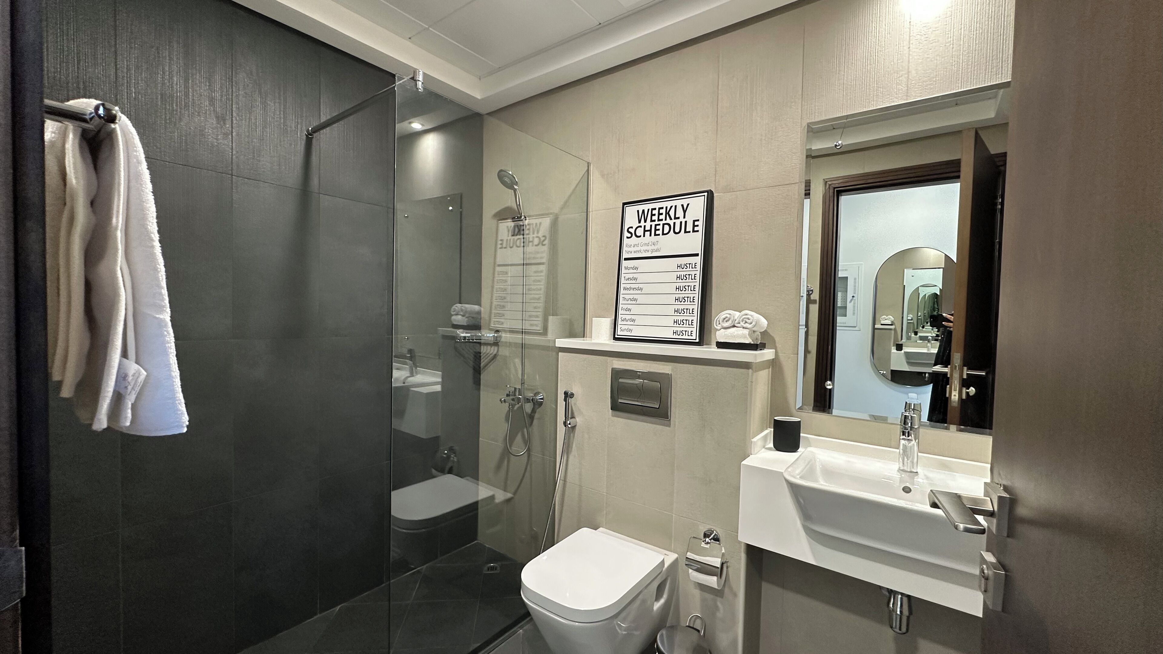 A modern bathroom with a glass shower enclosure, a sink with a mirror, and a weekly schedule sign.