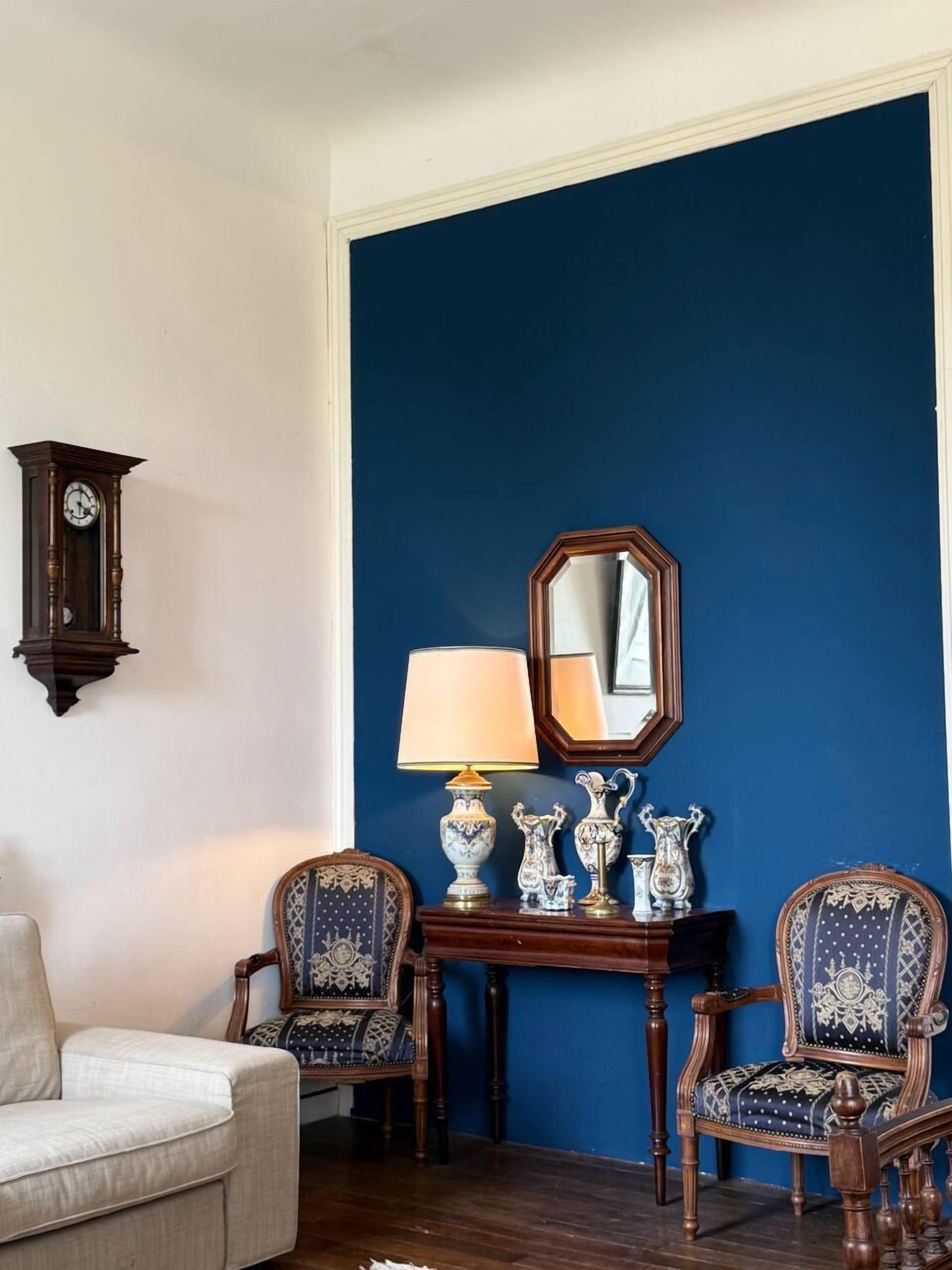 A room with a blue accent wall, a wooden side table with a lamp, two patterned chairs, a white sofa, and a wall-mounted clock.