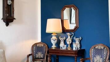 A room with a blue accent wall, a wooden side table with a lamp, two patterned chairs, a white sofa, and a wall-mounted clock.