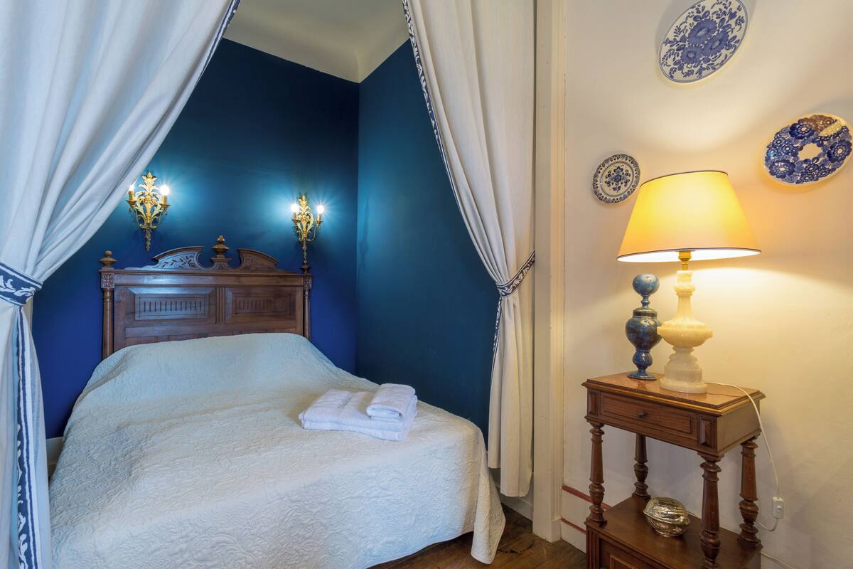 Una cama bien tendida con un edredón blanco, una mesita de noche con una lámpara y platos decorativos azules en la pared.