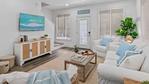 A modern living room with a white sofa set, a coffee table, a TV mounted on the wall, and a staircase leading to another level. - Seagrove Gem | 4 Bikes | Golf Cart | Yard Games (Santa Rosa Beach)