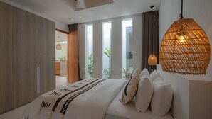 A modern bedroom with a large bed, a wooden wardrobe, and a view of the outdoors through large windows. - Modern 3 bedrooms pool villa near the beach, shops, and restaurants (Bali)