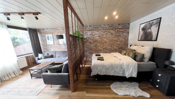 A modern bedroom with a brick wall, a large bed with a white bedspread, a grey sofa, and a wooden coffee table.