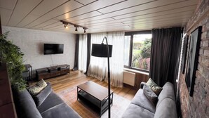 A modern living room with a flat-screen TV, a sofa, a coffee table, and a large window with curtains.