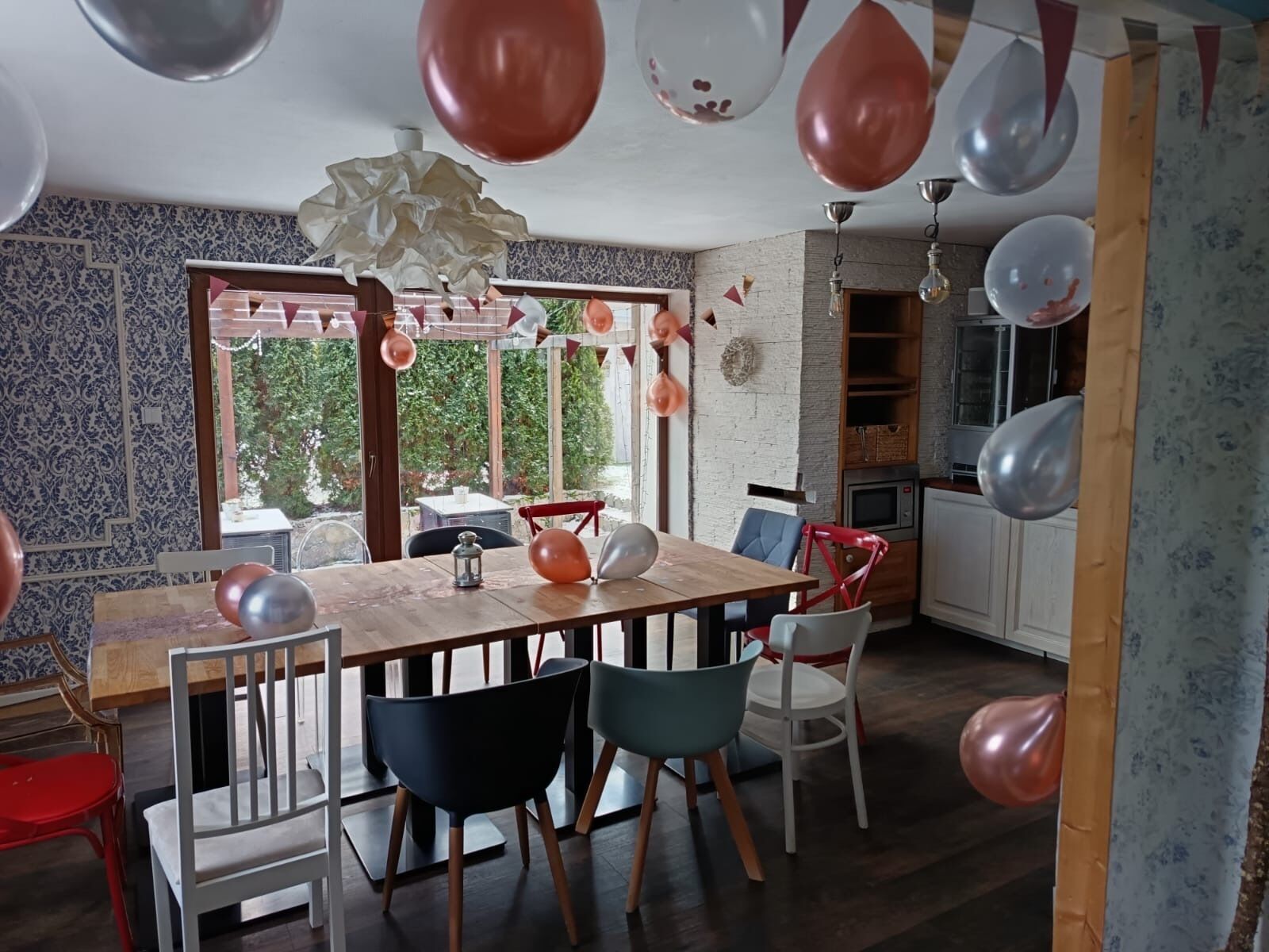 Een eetgedeelte met een houten tafel, stoelen en ballonnen. Er is een raam met uitzicht op groen.