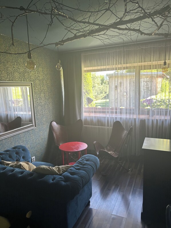 A room with a blue sofa, a small red table, and a dining table. There is a window with sheer curtains, and a mirror on the wall.