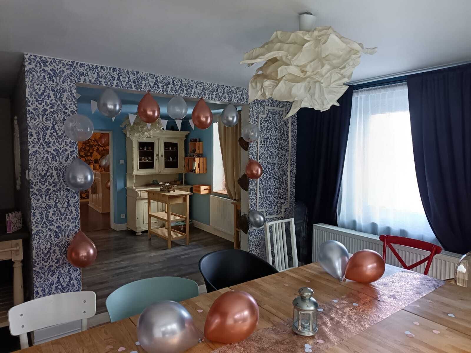 Et spiseområde med et træbord, stole og balloner. I baggrunden er der et køkken med skabe og en radiator.
