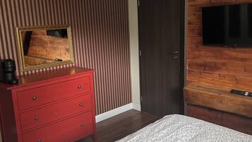 Un dormitorio con piso de madera, cómoda roja, espejo grande, colcha blanca estampada con flores, candelabro moderno y televisor de pantalla plana montado en una pared de madera.