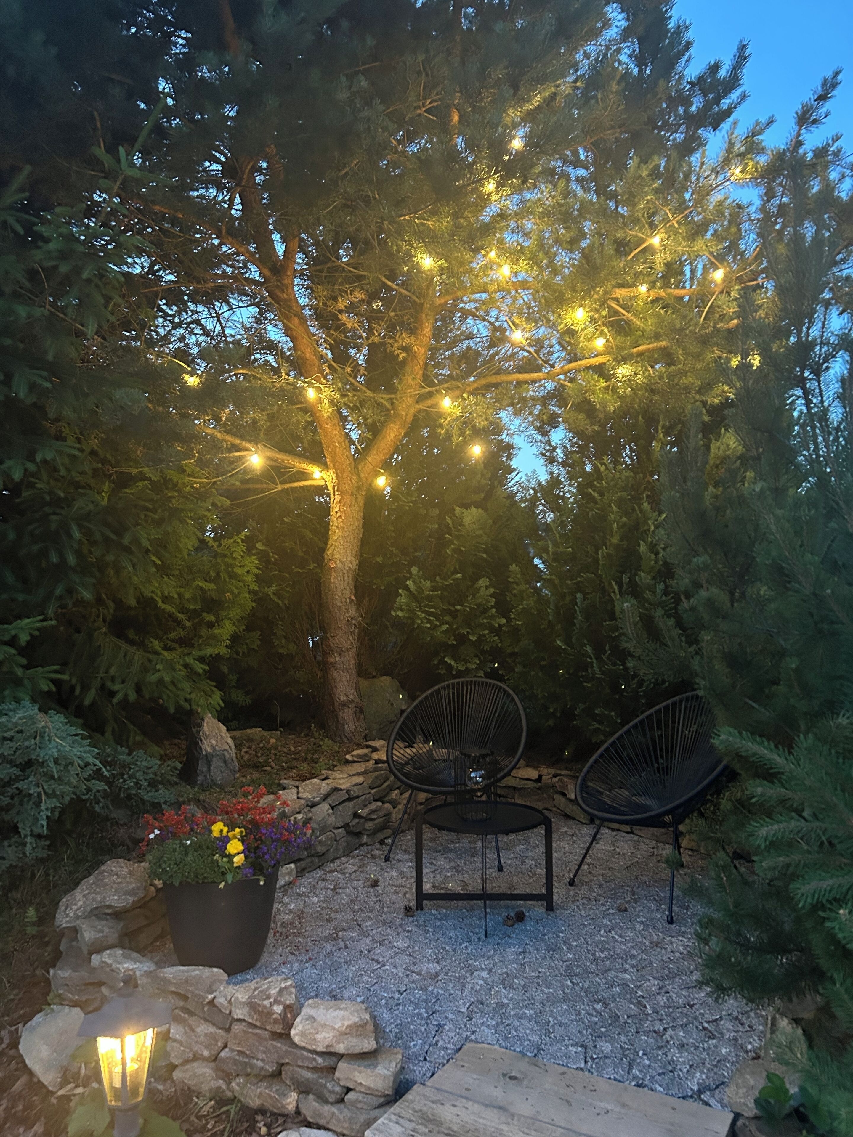 En uteplats med två svarta stolar, ett bord och ljusslingor som hänger på ett träd.