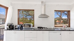A modern kitchen with white cabinets, a microwave, a stove, and a dishwasher. There are two windows with a view of trees and a building. - Kozyguru | Thornleigh | Townhouses (Thornleigh)